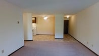 This single-floor unit presents an open-concept layout that seamlessly connects the living area to the kitchen and entryway. The living space is finished with neutral-toned carpet, contributing to a warm and understated atmosphere. Structural columns partially divide the interior, adding definition between zones while maintaining an open feel throughout.

The kitchen, visible from the main living area, features tile-style flooring, a white range, and dark wood cabinetry above. This practical layout allows for easy interaction between the kitchen and living spaces. A ceiling fan with a light fixture is centrally positioned, providing both air circulation and ambient lighting to the main area.

Near the kitchen, a dark-paneled entry door marks the unit's entrance, accompanied by a wall-mounted light fixture. The neutral beige and white wall tones throughout the space create a clean, move-in-ready canvas. Dark baseboards provide a subtle contrast against the light walls and flooring. Overall, the unit offers a functional, straightforward layout suited for everyday living.