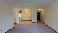 This single-floor unit presents an open-concept layout that seamlessly connects the living area to the kitchen and entryway. The living space is finished with neutral-toned carpet, contributing to a warm and understated atmosphere. Structural columns partially divide the interior, adding definition between zones while maintaining an open feel throughout.

The kitchen, visible from the main living area, features tile-style flooring, a white range, and dark wood cabinetry above. This practical layout allows for easy interaction between the kitchen and living spaces. A ceiling fan with a light fixture is centrally positioned, providing both air circulation and ambient lighting to the main area.

Near the kitchen, a dark-paneled entry door marks the unit's entrance, accompanied by a wall-mounted light fixture. The neutral beige and white wall tones throughout the space create a clean, move-in-ready canvas. Dark baseboards provide a subtle contrast against the light walls and flooring. Overall, the unit offers a functional, straightforward layout suited for everyday living.