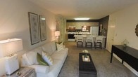 This single-floor residence showcases an open-concept layout that seamlessly connects the living room and kitchen, creating a spacious and functional environment. The living room is anchored by a plush white sofa dressed with green and yellow accent pillows, complementing the neutral-toned carpeting that extends throughout the space. A dark rectangular coffee table with a decorative floral centerpiece serves as the focal point of the seating area, while framed botanical artwork adorns the warm-toned walls, adding a refined touch to the room's aesthetic.

The kitchen is positioned toward the rear of the open floor plan and features light wood cabinetry, a white refrigerator, and a gas range with an overhead range hood. Dark accent walls in the kitchen create a striking contrast against the lighter cabinetry and appliances. A breakfast bar with seating for multiple guests provides a convenient dining option, with high stools lined along its edge. The kitchen also includes a sink positioned along the bar counter, making it both practical and accessible. Overall, the space presents a cohesive, inviting atmosphere enhanced by warm lighting and thoughtful décor choices.