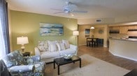 This single-floor residence offers a bright and inviting open-concept layout that seamlessly connects the living and dining areas. The living room is a comfortable and well-appointed space, anchored by a plush white sofa adorned with patterned throw pillows, a dark-finished coffee table, and a soft area rug. A ceiling fan overhead provides practical comfort, while a textured ceiling adds subtle dimension to the room. The walls are painted in a warm sage green, lending the space a calm and refreshing ambiance. A large landscape painting serves as a focal point on the main wall, complementing the nature-inspired color palette. A floor lamp provides warm, ambient lighting that enhances the cozy atmosphere. The space flows naturally into the dining area, which features a dark wood dining table and bar-height seating beneath a pendant light fixture. Decorative wall art and accent pieces add personality and charm to the dining nook. Durable wood-look flooring runs through the dining area, creating a cohesive transition between spaces. The overall design balances functionality and comfort, making this a practical yet aesthetically pleasing home suited for everyday living.