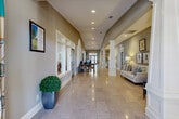 Located in Daphne, Alabama, this single-story community facility at 8254 CR-64 offers a thoughtfully designed and elegantly appointed interior that balances comfort with functionality. The expansive main corridor sets the tone upon entry, featuring gleaming polished tile flooring, tall ceilings, white decorative columns, and recessed lighting that illuminates the generous length of the hall. Comfortable seating and lush greenery accent the corridor, creating a welcoming first impression.

The main lounge area is a standout gathering space, furnished with upholstered armchairs, circular wood accent tables, and coastal-inspired artwork in soft aqua and gold tones. Rich hardwood flooring, crown molding, and large windows inviting natural light complete the relaxed, refined atmosphere.

A fully equipped kitchen features dark espresso cabinetry, stainless steel appliances including a refrigerator, dishwasher, and microwave, all set atop warm hardwood floors — ideal for community events and catering needs.

The fitness center is bright and well-lit, with multiple windows providing natural light and a selection of cardio and strength equipment including treadmills, stationary bikes, and a rowing machine.

Additional spaces include a sun-filled sitting room with garden views, an elegant conference room with a substantial trestle table and vaulted ceiling, and an intimate dining nook adorned with botanical artwork and classic furnishings.