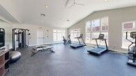 Located at 5 Kathryn Way in Middleborough, MA, this fitness center is a thoughtfully designed exercise facility offering a comprehensive workout environment on a single floor. The space is defined by striking vaulted ceilings that amplify the sense of openness, complemented by recessed lighting and a ceiling fan that ensure comfort during workouts. Large, multi-pane windows line the walls, flooding the interior with natural light and providing views of the surrounding community.

The flooring throughout is durable speckled rubber, ideal for high-traffic fitness use. Cardio equipment is well represented, with multiple treadmills, an elliptical trainer, and a stationary bike positioned to take full advantage of the window views. A multi-station cable machine anchors the strength training area, accompanied by a full rack of kettlebells, medicine balls, and foam rollers for functional fitness. Adjustable weight benches and dumbbells round out the free weights section.

A large wall mirror expands the visual depth of the space, while a wall-mounted flat-screen television adds an entertainment element. Glass double doors connect the fitness center to an adjacent area, enhancing the sense of flow and accessibility. The clean, neutral color palette of white and light gray walls contributes to a bright, motivating atmosphere throughout.