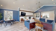 Located at 8210 Lakeshore Trail East Dr in Indianapolis, IN, this single-floor community clubhouse offers a well-appointed, open-concept layout that seamlessly blends a living room and kitchen into one cohesive gathering space. The living area is defined by striking vaulted ceilings with a cathedral peak, bold navy blue accent walls, and light hardwood-style flooring that runs throughout. A white brick fireplace with a mounted television serves as a focal point, while a large arched window and multiple French doors flood the space with natural light and provide views of the surrounding outdoor area. A ceiling fan adds both function and charm to the airy atmosphere. Comfortable navy blue sofas and lounge chairs are arranged around wooden accent tables, creating an inviting setting for relaxation or socializing. Adjacent to the living space, the kitchen features dark cabinetry with white quartz countertops, recessed lighting, a sink, and under-counter appliances. Open shelving displays decorative pieces, and a peninsula bar with white modern bar stools provides additional seating. Large windows bring in natural light, enhancing the clean, contemporary aesthetic. Interior windows also offer views into a fitness area, reinforcing this space's role as a full-service community amenity hub.