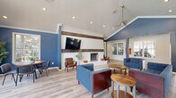 Located at 8210 Lakeshore Trail East Dr in Indianapolis, IN, this single-floor community clubhouse offers a well-appointed, open-concept layout that seamlessly blends a living room and kitchen into one cohesive gathering space. The living area is defined by striking vaulted ceilings with a cathedral peak, bold navy blue accent walls, and light hardwood-style flooring that runs throughout. A white brick fireplace with a mounted television serves as a focal point, while a large arched window and multiple French doors flood the space with natural light and provide views of the surrounding outdoor area. A ceiling fan adds both function and charm to the airy atmosphere. Comfortable navy blue sofas and lounge chairs are arranged around wooden accent tables, creating an inviting setting for relaxation or socializing. Adjacent to the living space, the kitchen features dark cabinetry with white quartz countertops, recessed lighting, a sink, and under-counter appliances. Open shelving displays decorative pieces, and a peninsula bar with white modern bar stools provides additional seating. Large windows bring in natural light, enhancing the clean, contemporary aesthetic. Interior windows also offer views into a fitness area, reinforcing this space's role as a full-service community amenity hub.