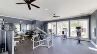 Located at 14411 Calloway Gap Rd in Raleigh, NC, this dedicated fitness center offers a well-appointed workout environment designed to support a comprehensive exercise routine. The single-floor space features a durable rubber-composite flooring in a neutral gray tone, providing both safety and comfort underfoot during workouts. The walls are finished in a deep slate blue, lending the space a focused and energizing atmosphere, while crisp white crown molding and a white ceiling add a clean, polished contrast. Natural light floods the room through an array of large, multi-pane windows along the exterior wall, offering pleasant views of the surrounding greenery and keeping the environment bright and inviting. Two ceiling fans are installed overhead, ensuring consistent air circulation throughout the space. The fitness area is equipped with a variety of strength-training machines, including a multi-station cable machine and several selectorized units, alongside a well-stocked dumbbell rack with an accompanying flat bench. Full-length mirrors line one wall, allowing users to monitor their form during exercises. The layout is thoughtfully organized to provide ample space between equipment, ensuring a safe and comfortable workout experience for all users.