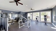 Located at 14411 Calloway Gap Rd in Raleigh, NC, this dedicated fitness center offers a well-appointed workout environment designed to support a comprehensive exercise routine. The single-floor space features a durable rubber-composite flooring in a neutral gray tone, providing both safety and comfort underfoot during workouts. The walls are finished in a deep slate blue, lending the space a focused and energizing atmosphere, while crisp white crown molding and a white ceiling add a clean, polished contrast. Natural light floods the room through an array of large, multi-pane windows along the exterior wall, offering pleasant views of the surrounding greenery and keeping the environment bright and inviting. Two ceiling fans are installed overhead, ensuring consistent air circulation throughout the space. The fitness area is equipped with a variety of strength-training machines, including a multi-station cable machine and several selectorized units, alongside a well-stocked dumbbell rack with an accompanying flat bench. Full-length mirrors line one wall, allowing users to monitor their form during exercises. The layout is thoughtfully organized to provide ample space between equipment, ensuring a safe and comfortable workout experience for all users.