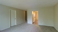 This two-floor residential space presents a practical and understated design throughout. The main living area is finished with wall-to-wall carpet in a light, neutral tone, contributing to a quiet and comfortable atmosphere. Walls are painted in a soft beige, creating a warm, cohesive backdrop across the space. An open passageway connects the living area to the kitchen, which features hard-surface flooring—a practical contrast to the carpeted surroundings. The kitchen is equipped with a full-sized refrigerator and appears to offer modest counter and storage space. A doorway visible from the living area leads to additional rooms, suggesting a functional layout designed for ease of movement between spaces. The overall aesthetic is clean and minimal, with simple white trim and standard fixtures throughout. The two-floor configuration provides additional living or sleeping areas beyond what is visible, expanding the overall footprint of the space. Natural light is present, lending brightness to the interior. The space is well-suited for those seeking a straightforward, move-in-ready environment with a neutral palette that accommodates a variety of furnishing styles.