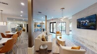 Located at 2900 N Braeswood Blvd in Houston, Texas, this sophisticated single-floor community amenity space is designed with a refined, contemporary aesthetic. The open-concept lounge area features herringbone-patterned wood-look tile flooring throughout, creating a warm yet polished foundation for the entire space. Plush cream sofas and accent chairs are arranged around a central coffee table, anchored by a large wall-mounted television set against a striking stone-clad accent wall. A linear pendant light fixture adds a modern architectural touch overhead, while floor-to-ceiling glass doors flood the space with natural light and provide access to an outdoor area.

The dining zone offers multiple seating arrangements with white marble-top tables and vibrant burnt-orange upholstered chairs on gold-tipped legs. Backlit vertical stone panels and a warm drum pendant light lend the dining area an elevated, restaurant-quality ambiance.

The kitchenette is a standout feature, showcasing powder-blue cabinetry paired with a shimmering gold hexagonal tile backsplash, marble countertops, open walnut shelving, a gold-accented faucet, and stainless steel appliances including a dishwasher and built-in refrigerator. A circular gold pendant light completes the space with a luxurious finishing touch.