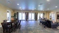 This single-floor gathering space is designed to accommodate a variety of social and functional uses within a warm, elegantly appointed interior. The open main room is generously proportioned, offering ample space for both dining and lounging areas. A large dining table with upholstered chairs anchors one side of the room, while a seating arrangement with a piano-style accent piece occupies the opposite end, creating a balanced and inviting layout.

The flooring features large-format stone-look tile in earthy tones, complemented by a decorative area rug that adds warmth and definition to the space. Recessed lighting is evenly distributed across the ceiling, providing consistent, well-diffused illumination throughout. The neutral-toned walls are dressed with floor-length drape panels in a rich charcoal gray, adding a refined, tailored look.

Three sets of French doors span the far wall, each framed by curtains and adorned with topiary-style potted plants, creating a seamless visual connection to the outdoor area beyond. Natural light pours in generously through these doors, enhancing the airy ambiance of the room. Decorative greenery and seasonal accents are thoughtfully placed throughout, contributing to the space's inviting and polished character.