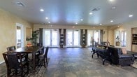 This single-floor gathering space is designed to accommodate a variety of social and functional uses within a warm, elegantly appointed interior. The open main room is generously proportioned, offering ample space for both dining and lounging areas. A large dining table with upholstered chairs anchors one side of the room, while a seating arrangement with a piano-style accent piece occupies the opposite end, creating a balanced and inviting layout.

The flooring features large-format stone-look tile in earthy tones, complemented by a decorative area rug that adds warmth and definition to the space. Recessed lighting is evenly distributed across the ceiling, providing consistent, well-diffused illumination throughout. The neutral-toned walls are dressed with floor-length drape panels in a rich charcoal gray, adding a refined, tailored look.

Three sets of French doors span the far wall, each framed by curtains and adorned with topiary-style potted plants, creating a seamless visual connection to the outdoor area beyond. Natural light pours in generously through these doors, enhancing the airy ambiance of the room. Decorative greenery and seasonal accents are thoughtfully placed throughout, contributing to the space's inviting and polished character.