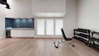 This dedicated fitness space is thoughtfully designed to support a wide range of workout routines in a clean, modern environment. The room features light wood-look flooring throughout, crisp white walls, and elevated ceilings that lend an open, airy feel to the space. Large windows fitted with horizontal blinds allow abundant natural light to filter in, while a ceiling fan ensures comfortable air circulation during workouts.

The cardio zone is equipped with a treadmill, upright stationary bike, and an elliptical trainer, complemented by a wall-mounted flat-screen television for entertainment during exercise. A dedicated strength area includes a dumbbell and kettlebell rack stocked with a varied selection of weights, along with an adjustable workout bench for versatile strength training.

A functional training station features a multi-use rack loaded with resistance bands, TRX suspension straps, and an assortment of medicine balls and stability balls in multiple sizes. Foam rollers are also on hand for recovery and stretching. Exposed ductwork above the functional training area adds an industrial design contrast to the otherwise polished interior.

A built-in cabinetry unit with a countertop and striking navy blue subway tile backsplash provides a practical refreshment or storage area. A side door offers convenient access, rounding out this comprehensive and well-appointed fitness facility.