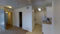 This single-story space presents a straightforward, functional layout with clearly defined living and kitchen areas. The living space features wall-to-wall carpeting and includes a large sliding-door closet, offering generous built-in storage with visible shelving inside. Neutral white walls throughout contribute to a clean, understated aesthetic, while the textured popcorn ceiling adds a classic residential character.

Adjacent to the living area, the kitchen is finished with light-toned vinyl tile flooring, providing an easy transition between spaces. White cabinetry lines both the upper and lower sections of the kitchen walls, offering ample storage and a cohesive look. The kitchen is equipped with a white electric coil-top range and a full-size white refrigerator, with countertops providing functional preparation space. A ceiling-mounted fluorescent light fixture provides direct illumination over the kitchen workspace.

Ventilation is served by ceiling-mounted HVAC registers visible throughout the unit, ensuring consistent climate control. The overall palette of whites and neutral tones gives the space a simple, practical character well-suited for everyday living.