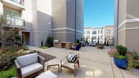 This well-appointed amenity space seamlessly blends indoor comfort with outdoor living across a single level. The interior opens into a sophisticated lounge area finished with wide-plank hardwood-style flooring, recessed lighting, and large windows dressed with elegant sheer curtains that invite abundant natural light. A standout feature is the gourmet kitchen, highlighted by an expansive marble-topped island with seating for multiple guests, dark cabinetry, a dramatic marble backsplash, and a striking gold-toned linear chandelier that adds a refined touch overhead. Adjacent to the kitchen, a dedicated dining nook offers restaurant-style leather banquette booths separated by a central divider, each booth accompanied by a wall-mounted TV and illuminated by decorative globe pendant lighting — creating an intimate yet social atmosphere. The interior transitions seamlessly to the outdoors through glass double doors, leading to a generous open-air courtyard. The patio features a built-in brick grilling station, lush planters with seasonal greenery, and multiple seating arrangements with wicker and upholstered furniture set against a backdrop of a multi-story residential building. The overall design balances modern elegance with functional communal living, making it an ideal gathering space.