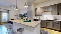 This well-appointed single-floor residence offers a thoughtfully designed open-concept layout that seamlessly connects the kitchen, dining, and living areas. The kitchen is a standout feature, showcasing rich dark-toned cabinetry, granite countertops, a large central island with pendant lighting above, and a full suite of stainless steel appliances including a range, microwave, and dishwasher. Light wood-style flooring flows throughout, complementing the bright, white walls and elevated ceilings that contribute to an airy, spacious feel. The living room is anchored by glass French doors that invite natural light and provide outdoor access, while a ceiling fan and stylish accent lighting enhance the room's comfort and ambiance. The bedroom is generously proportioned, featuring a large window with views to the outside, light wood flooring, and access to a spacious walk-in closet outfitted with built-in shelving, hanging rods, and dedicated storage sections. The bathroom is clean and functional, featuring dark wood cabinetry paired with granite countertops, a sink vanity with a mirror, and a full tub-shower combination with tile surround. A dedicated laundry room with a washer and dryer unit, open shelving, and additional storage space rounds out this practical and stylishly finished residence.