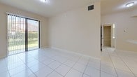This well-appointed single-floor unit offers a clean, open layout finished throughout with large-format white ceramic tile flooring that enhances the sense of brightness and space. The kitchen is a standout feature, thoughtfully designed with floor-to-ceiling wood-grain cabinetry in a warm gray-brown tone, complemented by a marble-look tile backsplash and light countertops. It includes ample upper and lower cabinet storage, a stainless steel sink, and white appliances — including a refrigerator, electric range, over-the-range microwave, and dishwasher — making it both functional and visually cohesive. A peninsula counter provides additional workspace and separates the kitchen from the adjoining living area. The living space is generously sized with neutral beige walls and a sliding glass door that fills the room with natural light and provides direct access to a greenery-filled outdoor area. The bathroom is finished entirely in white, featuring large wall tiles in the tub/shower surround, a built-in vanity with under-sink storage, and a bright window with plantation shutters. A louvered closet door adjacent to the bathroom provides convenient storage access, completing this clean, move-in ready space.