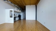 This single-floor loft showcases a striking industrial aesthetic defined by soaring ceilings with warm wood paneling and exposed metal ductwork, creating a bold architectural statement. Slim black structural columns punctuate the open main living area, reinforcing the industrial character while complementing the clean white walls throughout. The expansive main area features smooth hardwood flooring in a warm honey tone, lending a welcoming contrast to the raw industrial overhead elements.

The kitchen is integrated into the space via a raised platform, partially separated by a half-wall that defines the area without fully enclosing it. It is equipped with stainless steel appliances, including a range and refrigerator, and features white cabinetry with ample storage. Pendant lights hang above the kitchen area, providing focused task lighting that complements the industrial-style overhead fixtures in the main space.

The overall layout is open and versatile, offering generous room to configure living and dining areas as desired. The combination of exposed materials, high ceilings, and unobstructed floor space gives the unit a spacious, loft-style ambiance suited for a variety of uses.
