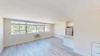 This single-floor unit presents a clean, contemporary design characterized by an open-concept layout and a bright, airy atmosphere. Light-toned hardwood flooring runs throughout the space, complementing the crisp white walls and creating a cohesive, modern aesthetic.

The main living area is generously proportioned, anchored by a wide bank of windows fitted with horizontal blinds that frame sweeping views of surrounding buildings and a verdant hillside. A wall-mounted HVAC unit provides climate control, while abundant natural light fills the room throughout the day.

The kitchen is positioned adjacent to the living area and is defined by high-gloss white cabinetry extending to the ceiling, recessed lighting, stainless steel appliances including a refrigerator and dishwasher, and a marble-veined quartz peninsula that doubles as a breakfast bar and extends into the living space. A sleek undermount sink and matching backsplash complete the refined look.

The bathroom maintains the unit's consistent white palette, featuring light wood-toned flooring, a floating white vanity with a quartz countertop, a mirrored medicine cabinet, and recessed ceiling lighting. The layout is clean and functional, with a walk-in shower area and chrome fixtures throughout.