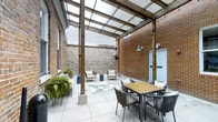 This inviting courtyard space offers a seamless blend of historic architectural character and contemporary outdoor design. The space is defined by striking exposed brick walls — a mix of warm red brick and rustic stone — that lend a sense of heritage and texture to the environment. Overhead, a pergola structure fitted with translucent polycarbonate panels diffuses natural light evenly throughout the space, providing shelter while maintaining a bright, airy atmosphere regardless of the weather.

The tiled floor, laid in large neutral-toned pavers, provides a clean and polished foundation that complements the raw, industrial character of the surrounding walls. The courtyard is thoughtfully arranged with distinct zones for dining and lounging. A slatted wood-top dining table surrounded by woven rope chairs anchors one area, while a separate seating arrangement featuring cushioned chairs and a decorative area rug creates a relaxed lounge corner. Lush potted ferns and hanging planters introduce organic greenery, softening the hardscape and adding life to the setting.

Wall-mounted industrial-style sconce lighting and concrete support posts reinforce the space's architectural integrity. A subtle door set into the brick wall provides discreet access to the surrounding structure. Overall, the courtyard exudes a refined yet relaxed ambiance, ideal for both casual gatherings and quiet outdoor enjoyment.