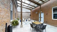 This inviting courtyard space offers a seamless blend of historic architectural character and contemporary outdoor design. The space is defined by striking exposed brick walls — a mix of warm red brick and rustic stone — that lend a sense of heritage and texture to the environment. Overhead, a pergola structure fitted with translucent polycarbonate panels diffuses natural light evenly throughout the space, providing shelter while maintaining a bright, airy atmosphere regardless of the weather.

The tiled floor, laid in large neutral-toned pavers, provides a clean and polished foundation that complements the raw, industrial character of the surrounding walls. The courtyard is thoughtfully arranged with distinct zones for dining and lounging. A slatted wood-top dining table surrounded by woven rope chairs anchors one area, while a separate seating arrangement featuring cushioned chairs and a decorative area rug creates a relaxed lounge corner. Lush potted ferns and hanging planters introduce organic greenery, softening the hardscape and adding life to the setting.

Wall-mounted industrial-style sconce lighting and concrete support posts reinforce the space's architectural integrity. A subtle door set into the brick wall provides discreet access to the surrounding structure. Overall, the courtyard exudes a refined yet relaxed ambiance, ideal for both casual gatherings and quiet outdoor enjoyment.