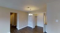 This single-floor space presents a clean, well-maintained interior defined by warm beige walls and rich, dark hardwood-style flooring that runs throughout. The main living area is illuminated by a stylish multi-bulb pendant chandelier, which serves as a focal point and adds a touch of elegance to the room. Overhead, a flush ceiling vent ensures efficient climate control throughout the space.

The layout opens to several adjoining areas, accessible through white panel doors that contribute to the bright, cohesive aesthetic. A closet with open bifold doors reveals a dedicated storage area with a built-in shelf, ideal for organized storage. An additional doorway leads to what appears to be a secondary room or hallway, enhancing the functional flow of the floor plan. To the right, a partial view of granite-style countertop suggests proximity to a kitchen or utility area.

White baseboards frame the perimeter of the space, adding a crisp finishing detail that complements the neutral palette. Overall, the space conveys a tidy, move-in-ready atmosphere with practical storage solutions and a warm, inviting ambiance suitable for everyday living.