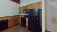 Located at 111 W Marshall in Richmond, Virginia's 23220 zip code, this two-floor unit blends industrial character with functional living. The space is distinguished by richly stained concrete floors throughout, lending a bold, urban aesthetic that ties the interior together cohesively. The kitchen is well-appointed with warm wood cabinetry extending to the ceiling for ample storage, complemented by black appliances including a refrigerator, electric range with over-the-range microwave, and a sink with under-cabinet storage. The bathroom features a full tub-shower combination with clean white surround panels, a toilet, and vanity with mirrored medicine cabinet above. An open area provides versatile living or sleeping space with neutral white walls and clean baseboard detailing. The staircase connecting the two floors features a contemporary metal railing with horizontal balusters and wood accents, leading to an upper level where exposed concrete structural columns and visible industrial piping along the ceiling add a distinctive loft-like character. Red fire suppression lines and galvanized ductwork overhead reinforce the building's converted industrial heritage, creating a uniquely textured and characterful living environment in the heart of Richmond.