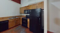 Located at 111 W Marshall in Richmond, Virginia's 23220 zip code, this two-floor unit blends industrial character with functional living. The space is distinguished by richly stained concrete floors throughout, lending a bold, urban aesthetic that ties the interior together cohesively. The kitchen is well-appointed with warm wood cabinetry extending to the ceiling for ample storage, complemented by black appliances including a refrigerator, electric range with over-the-range microwave, and a sink with under-cabinet storage. The bathroom features a full tub-shower combination with clean white surround panels, a toilet, and vanity with mirrored medicine cabinet above. An open area provides versatile living or sleeping space with neutral white walls and clean baseboard detailing. The staircase connecting the two floors features a contemporary metal railing with horizontal balusters and wood accents, leading to an upper level where exposed concrete structural columns and visible industrial piping along the ceiling add a distinctive loft-like character. Red fire suppression lines and galvanized ductwork overhead reinforce the building's converted industrial heritage, creating a uniquely textured and characterful living environment in the heart of Richmond.