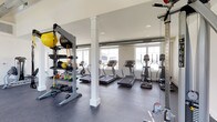 This fitness center offers a comprehensive workout environment thoughtfully organized across a single open floor. The space is characterized by high ceilings — including a striking clerestory section with skylights — exposed ductwork, and large windows that flood the interior with natural light and provide views of the surrounding outdoors, including glimpses of a pool area.

The cardio zone features a row of treadmills, elliptical trainers, and a stationary bike, all positioned along the windowed wall to maximize natural light during workouts. A dedicated strength training area houses multi-station cable machines and a full dumbbell rack with a wide range of weights. Functional training needs are met with a well-stocked free-standing rack loaded with medicine balls, resistance bands, kettlebells, and suspension training straps.

The flooring throughout is durable rubber gym matting, providing safety and comfort underfoot. White structural columns punctuate the space, complementing the clean, neutral color palette of white walls and light ceilings. Recessed lighting supplements the ample natural light. The overall ambiance is bright, clean, and functional — designed to support a wide range of fitness activities in a welcoming and organized environment.