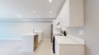 This well-appointed single-floor unit offers a thoughtfully designed layout that maximizes every square foot across one bedroom, one bathroom, and a combined kitchen and living area. The kitchen is a standout feature, showcasing high-gloss white cabinetry, bright quartz countertops, a large island with an integrated sink, and a full suite of stainless steel appliances including a range, dishwasher, and refrigerator. Recessed lighting illuminates the space, complementing the light neutral tones and light-toned laminate flooring found throughout. The open-concept design allows the kitchen to flow naturally into the living area, which benefits from a wall-mounted mini-split climate control unit and direct access to an outdoor balcony or patio. Large windows in both the living and bedroom areas invite generous natural light and offer views of surrounding greenery. The bedroom is a cozy, well-proportioned retreat featuring a baseboard heater, soft grey walls, and the same warm flooring carried throughout the unit. The bathroom is finished in a clean, minimalist style with neutral tile flooring and chrome accents. Overall, the unit presents a modern, move-in-ready aesthetic with a cohesive palette of whites and soft greys.