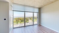 This single-floor high-rise unit is defined by its striking floor-to-ceiling windows that frame breathtaking panoramic views of a verdant park, mature trees, and a shimmering body of water beyond. The open living area benefits enormously from this wall of glass, flooding the space with natural light and creating a seamless visual connection to the outdoors. Durable wood-look flooring runs throughout the main living areas, complementing the unit's clean, contemporary aesthetic.

The galley-style kitchen is well-appointed with white upper cabinetry, gray lower cabinets, crisp white subway tile backsplash, and gleaming white countertops. Stainless steel appliances — including a range, microwave, dishwasher, and refrigerator — provide a polished, modern finish. A window at the end of the kitchen allows natural light to filter through.

The living space features defined alcoves along one wall, offering flexible arrangement options, while the large sliding glass door continues the theme of expansive views. Additional rooms include a carpeted bedroom or flex room with neutral tones, a bathroom with wood-look flooring, a dark-toned vanity, and clean white fixtures. The unit's exposed concrete ceiling elements lend an industrial character that contrasts elegantly with the otherwise refined, modern interior.