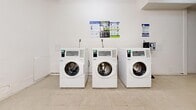 Located at 645 Northcliffe Blvd in Toronto's west end, this shared laundry facility offers a clean, functional, and well-organized space designed to serve building residents efficiently. The interior is finished with crisp white walls and durable light-toned tile flooring throughout, creating a bright and tidy environment illuminated by overhead fluorescent lighting.

The main laundry area houses a bank of commercial-grade front-loading washers — three Huebsch units positioned side by side — alongside a matching set of three top-load dryers, providing ample capacity for residents. The machines are equipped with digital payment interfaces, supporting convenient cashless transactions. Informational signage is posted on the walls, offering guidance on machine usage and payment options.

A freestanding utility sink provides additional functionality for hand-washing or pre-treating garments. A sturdy wooden folding table is centrally placed, offering a practical surface for sorting and folding laundry. A bulletin board mounted on the wall near the entrance serves as a communication hub for building notices.

The space connects to an adjacent hallway entry area, which features a fire extinguisher, waste bin, and access door, ensuring safety and ease of circulation. The overall layout is practical and efficiently organized, making this laundry facility a straightforward and accessible amenity for all building occupants.