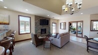 This single-floor community clubhouse is thoughtfully designed to offer residents a comfortable and functional gathering space. The main lounge area serves as the centerpiece of the facility, featuring a dramatic floor-to-ceiling stacked stone fireplace with a wall-mounted flat-screen television above it. The lounge is furnished with a plush sectional sofa and accent chairs arranged around a round coffee table, creating an inviting setting for relaxation or socializing. A vaulted ceiling detail and an elegant chandelier add architectural interest and a refined ambiance to the room. Multiple windows throughout the lounge allow natural light to fill the space, with views of an outdoor pool area visible from several vantage points. Adjacent to the lounge is a reception and gathering area, complete with an oval dining table, chairs, and a raised bar-height counter with bar stools — ideal for meetings or casual interactions. A service counter with organizational elements and direct access to the exterior makes this area functional for administrative purposes as well. The fitness center is well-appointed with cardio equipment including treadmills and an elliptical machine, all positioned near windows that provide ample natural light and outdoor views. Consistent warm-toned walls, dark wood trim, and carpeted flooring tie the spaces together with a cohesive, welcoming aesthetic throughout.