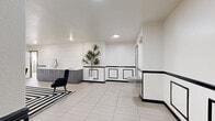 Located at 240 S Monaco St Pkwy in Denver, CO, this spa lounge offers a well-appointed, single-floor wellness retreat with a cohesive and striking black-and-white design throughout. The main lounge area serves as the centerpiece of the space, featuring white walls accented by bold black wainscoting with decorative panel detailing. Plush black accent chairs are arranged around a graphic black-and-white geometric area rug, while a large Roman numeral wall clock and decorative greenery add character and warmth to the setting. Recessed lighting illuminates the lounge evenly, creating a clean and inviting atmosphere. Adjacent to the lounge, a vanity area with gray cabinetry and a countertop mirror provides a functional grooming station. A dedicated changing or coat room with wall-mounted hooks connects seamlessly to the rest of the facility. The bathrooms include a classic wood-lined dry sauna with tiered benches and a built-in heater, offering an authentic sauna experience. A fully tiled steam room features floor-to-ceiling white tile, a mosaic tile floor in warm earth tones, and built-in bench seating. Multiple private shower stalls with frosted glass doors and mosaic tile flooring complete the bathing amenities, rounding out a comprehensive and thoughtfully designed spa lounge.