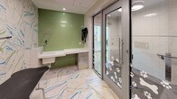 Located at 4850 Rugby Ave in Bethesda, Maryland, this purpose-built pet grooming facility is a single-floor space designed with both function and aesthetic appeal in mind. The room is immediately striking for its bold, artistic tile work — large-format floor and wall tiles feature an abstract white, blue, and green marbled pattern that lends the space a clean yet lively character. The walls transition between distinct zones, with vibrant teal subway tiles lining a spacious walk-in pet shower stall equipped with a professional-grade flexible spray hose and a built-in shelf. An adjacent washing station features a raised white tub with its own retractable sprayer, set against a backdrop of sage green glossy tile. A built-in under-counter storage unit and wall-mounted drying accessories complete the grooming station. The floors carry the vivid marbled tile throughout, maintaining design cohesion across the space. Entry is provided through sleek glass sliding doors etched with playful paw print and bone motifs, offering a welcoming and pet-friendly aesthetic. Recessed and flush-mounted ceiling lighting ensures the space is well-lit throughout. Every detail reflects a thoughtfully curated grooming environment built for practicality and comfort.