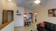 This single-floor residence offers an open-concept layout that seamlessly connects the living, dining, and kitchen areas into one cohesive, functional space. The living area is anchored by a ceiling fan with a light fixture, which enhances air circulation and provides ample overhead lighting. Light-toned walls and neutral flooring throughout contribute to a clean, airy atmosphere.

The kitchen is efficiently designed with light wood cabinetry, a range, and a raised breakfast bar that accommodates bar-height seating — ideal for casual dining or entertaining. Floating shelves on an adjacent accent wall provide decorative display space and add a modern touch to the open layout.

Conveniently positioned just off the main living area is an in-unit laundry alcove, housing a stacked washer and dryer — a practical feature that adds everyday convenience. A hallway leads to a bathroom outfitted with warm wood cabinetry and a well-lit vanity mirror, maintaining the consistent neutral aesthetic found throughout the space.

The overall design emphasizes openness and practicality, making efficient use of every area. The consistent color palette of warm neutrals and natural wood tones creates a welcoming and cohesive ambiance throughout the entire floor plan.