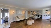 This thoughtfully designed single-floor space exudes a timeless elegance through its carefully curated aesthetic and well-appointed interiors. The living area serves as the heart of the space, showcasing rich hardwood flooring that runs throughout and adds warmth to the neutral, light gray walls. A classic camelback sofa in cream upholstery anchors the seating area, complemented by a wrought iron coffee table and a pair of tufted ottomans that add texture and charm. Tall arc floor lamps flank the room, providing ambient lighting that enhances the inviting atmosphere. A striking metallic wall art piece serves as a focal point, reflecting light and adding visual interest to the space. Beyond the living area, a dining or secondary lounge space is visible, featuring a fireplace with a classic black mantle, built-in shelving, and additional seating with elegant white chairs. The open layout allows for a natural flow between areas, making the space feel connected and spacious. Chrome and glass accent furniture lend a modern contrast to the more traditional design elements, resulting in a balanced and polished interior that is both functional and aesthetically refined.