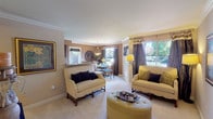This single-floor space presents an open-concept layout connecting the living and dining areas in a cohesive, well-appointed design. The living area is anchored by two cream-toned loveseats with dark wood legs, accented by navy and yellow decorative pillows, and centered around a tufted round ottoman that doubles as a coffee table. Crown molding runs along the ceiling, adding a refined architectural detail throughout the space.

Natural light flows generously through large sliding glass doors and wide windows dressed with rich chocolate brown curtains, offering views of lush greenery outside. A warm neutral color palette of tan and cream tones unifies the walls, carpet, and furnishings, creating a calm and inviting ambiance. A floor lamp with a yellow shade provides warm supplemental lighting.

The adjacent dining area, visible through a pass-through opening, features a round table with upholstered chairs beneath a classic pendant light, making it well-suited for intimate gatherings. Decorative wall art and a sunburst mirror add personality and visual interest throughout the space. Overall, the layout and styling suggest a comfortable, classic aesthetic with thoughtful attention to detail.