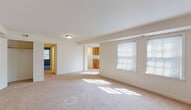 This single-floor residence offers a thoughtfully laid-out living space with a clean, neutral aesthetic throughout. The expansive living room is carpeted in a warm beige tone and is bathed in natural light streaming through multiple windows fitted with white horizontal blinds. Structural beams add subtle architectural definition to the open area, which flows seamlessly toward the kitchen through a wide pass-through opening.

The galley-style kitchen is well-appointed with an abundance of warm oak cabinetry on both upper and lower levels, providing ample storage. Grey laminate countertops run the length of the kitchen on both sides, complemented by a white electric range, a dishwasher, and light-toned tile flooring. A window above the sink area allows natural light to brighten the workspace.

The generously proportioned bedroom features the same plush beige carpet as the living areas and is illuminated by dual windows, creating an airy and comfortable environment. Closet access is readily available within the room.

The full bathroom is finished with crisp white tile flooring accented with blue diamond inlays, and the tub surround features a distinctive blue-and-white checkered tile border. A window above the tub provides natural ventilation, and a wood-toned vanity with a light countertop completes the space. Overall, the residence presents a clean, functional, and move-in ready environment.