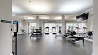 This fitness center offers a generously sized, single-level workout space designed to accommodate a comprehensive range of exercise equipment and training activities. The interior is finished in a clean, neutral palette with white walls, white structural columns, and speckled rubber flooring that provides both durability and comfort underfoot.

The space is divided into functional zones. The cardio area features multiple treadmills and elliptical machines positioned along a wall of large floor-to-ceiling windows, allowing natural light to fill the room and providing outdoor views of surrounding greenery. Ceiling fans and suspended linear fluorescent fixtures ensure consistent ventilation and illumination throughout.

The strength training section includes a dumbbell rack with a full range of free weights, adjustable benches, a multi-station cable machine, and additional selectorized strength equipment. Large wall-mounted mirrors expand the visual depth of the space and allow users to monitor their form.

A separate alcove toward the rear accommodates additional strength machines and is accented by a colorful wall-mounted artwork piece, adding a vibrant focal point. The entrance area features glass double doors, a sanitation station, and wall-mounted safety features. A wall-mounted television serves the main training floor. Overall, the space presents a bright, functional, and well-organized fitness environment.