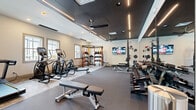 This modern fitness center is thoughtfully designed to support a wide range of exercise routines. The space is divided into two distinct zones: a main gym area and a dedicated yoga studio, separated by a sleek floor-to-ceiling glass partition with framed glass doors.

The main workout area features a striking slatted dark ceiling with linear LED lighting and ceiling fans, creating a dynamic yet comfortable environment. Large windows along the walls invite ample natural light throughout the space. The floor transitions between carpet and hardwood-style surfaces, delineating workout zones. Equipment includes Matrix-brand treadmills, ellipticals, and adjustable benches, alongside a multi-functional training rack with resistance attachments, a medicine ball station, and a tiered dumbbell rack offering a full range of free weights. A wall-mounted flat-screen TV adds an entertainment element to the workout experience. Full-length mirrors line the walls, enhancing the sense of space and allowing users to monitor their form.

The yoga studio is a bright, open room with light hardwood flooring, recessed lighting, ceiling fans, and large windows that flood the area with natural light. Spin bikes, a treadmill, and stability balls are also housed here, making it versatile for cardio and group fitness classes alike.