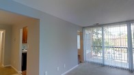 This single-floor space offers a straightforward and functional layout designed for comfortable living. The main living area is characterized by neutral gray-toned walls and light-colored carpeted flooring, creating a calm and understated atmosphere throughout. A large window fitted with vertical blinds dominates one wall, allowing an generous amount of natural light to filter into the room while offering views of lush greenery just outside. The window treatment provides both privacy control and flexibility in managing the brightness of the space. The ceiling is smooth and white, lending an airy, open feel to the interior. An open doorway or passageway connects the main living area to an adjacent room, which features warm-toned walls, suggesting a distinct yet complementary space — possibly a hallway or secondary room. Visible HVAC vents integrated into the ceiling indicate a central climate control system. The overall ambiance of the space is clean, bright, and move-in ready, with a minimalist aesthetic that allows residents to personalize the interior with ease. The connection to outdoor greenery visible through the window adds a pleasant natural element to the living environment.