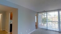 This single-floor space offers a straightforward and functional layout designed for comfortable living. The main living area is characterized by neutral gray-toned walls and light-colored carpeted flooring, creating a calm and understated atmosphere throughout. A large window fitted with vertical blinds dominates one wall, allowing an generous amount of natural light to filter into the room while offering views of lush greenery just outside. The window treatment provides both privacy control and flexibility in managing the brightness of the space. The ceiling is smooth and white, lending an airy, open feel to the interior. An open doorway or passageway connects the main living area to an adjacent room, which features warm-toned walls, suggesting a distinct yet complementary space — possibly a hallway or secondary room. Visible HVAC vents integrated into the ceiling indicate a central climate control system. The overall ambiance of the space is clean, bright, and move-in ready, with a minimalist aesthetic that allows residents to personalize the interior with ease. The connection to outdoor greenery visible through the window adds a pleasant natural element to the living environment.