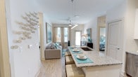 Located at 4320 Commons Dr W in Destin, Florida, this single-floor residence offers a thoughtfully designed open-concept layout that seamlessly connects the kitchen, dining, and living areas. The kitchen is anchored by a generous granite-topped island with bar seating, providing both a functional prep space and a natural gathering point. Pendant lighting above the island adds a polished touch, while the adjoining dining and living areas benefit from abundant natural light streaming through multiple windows dressed in elegant neutral drapery. The living space features smooth light wood-style flooring that extends throughout, enhancing the airy, cohesive feel of the interior. Vaulted ceilings with a ceiling fan lend an added sense of openness to the main living area. The coastal-inspired color palette of soft whites, warm beiges, and aqua accents creates a calm and inviting atmosphere throughout. A private bedroom is accessible directly from the main living area, offering a quiet retreat with its own tasteful décor. The overall design balances modern convenience with relaxed, beachy elegance — well-suited to the Destin coastal lifestyle.