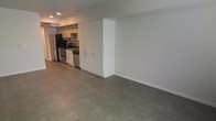 This single-floor space features an open-plan layout that seamlessly integrates the kitchen and living areas. The kitchen is equipped with stainless steel appliances, including a refrigerator, oven/range, and dishwasher, complemented by modern flat-front white cabinetry that extends floor to ceiling, providing ample storage. A dedicated range hood is installed above the cooktop, adding both functionality and a clean aesthetic. The cabinetry design creates a natural visual divider between the kitchen and the open living area without fully enclosing either space. Polished concrete flooring runs continuously throughout, lending an industrial yet refined character to the interior. The walls are finished in a neutral white, contributing to a bright and clean atmosphere. Recessed lighting is installed in the ceiling, ensuring consistent illumination across the space. The overall design is minimal and contemporary, with a practical, uncluttered layout that prioritizes open space and functionality. The neutral palette of white walls, grey floors, and stainless steel accents creates a cohesive and modern aesthetic throughout.