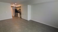 This single-floor space features an open-plan layout that seamlessly integrates the kitchen and living areas. The kitchen is equipped with stainless steel appliances, including a refrigerator, oven/range, and dishwasher, complemented by modern flat-front white cabinetry that extends floor to ceiling, providing ample storage. A dedicated range hood is installed above the cooktop, adding both functionality and a clean aesthetic. The cabinetry design creates a natural visual divider between the kitchen and the open living area without fully enclosing either space. Polished concrete flooring runs continuously throughout, lending an industrial yet refined character to the interior. The walls are finished in a neutral white, contributing to a bright and clean atmosphere. Recessed lighting is installed in the ceiling, ensuring consistent illumination across the space. The overall design is minimal and contemporary, with a practical, uncluttered layout that prioritizes open space and functionality. The neutral palette of white walls, grey floors, and stainless steel accents creates a cohesive and modern aesthetic throughout.