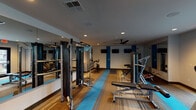 Located at 4410 Marburg Ave in Cincinnati, Ohio, this single-floor fitness center is designed to support a comprehensive workout experience. The space is laid out to accommodate both strength training and cardiovascular exercise, with distinct zones dedicated to each. The flooring features a combination of wood-tone carpeting and blue runner accents that guide movement through the facility and add a modern visual element to the overall design.

The strength training area is well-equipped with a cable machine, weight bench, and additional resistance equipment. Large mirrors line one wall, enhancing the sense of openness and allowing users to monitor their form. Blue accent columns punctuate the walls, reinforcing a clean, contemporary aesthetic throughout. Mounted flat-screen televisions are positioned for easy viewing from multiple workout stations, and a ceiling fan provides additional ventilation.

The cardio section is situated near a bank of large windows fitted with blinds, allowing natural light to fill the area while maintaining privacy. Matrix-branded stationary bikes, a treadmill, and an elliptical trainer are arranged to take advantage of the natural light and outdoor views. A dedicated spin bike area with storage cubbies and wall hooks offers an organized space for cycling workouts. Recessed lighting throughout ensures consistent illumination, completing a well-appointed and functional fitness environment.