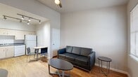 This deluxe efficiency studio is a thoughtfully organized single-floor unit that maximizes every inch of its compact footprint. The open-concept living area features warm hardwood-style flooring, neutral gray walls, and tall ceilings that lend a sense of openness. A large window allows natural light to fill the space, creating a bright and welcoming atmosphere. The living area comfortably accommodates a sofa and coffee table arrangement, while a ceiling fan adds practical comfort overhead.

The kitchen is well-equipped with light wood cabinetry, dark granite-style countertops, and a full suite of white appliances including a range with electric cooktop, over-the-range microwave, dishwasher, and a full-size refrigerator. Track lighting overhead ensures the kitchen is well-lit for everyday use. A half-wall partition subtly separates the kitchen from the sleeping area, which features carpeted flooring for added comfort.

The bathroom is clean and functional, complete with a tub-shower combination, a single vanity with matching cabinetry and dark countertop, an overhead storage cabinet, and hardwood-style flooring. The layout as a whole is efficient and cohesive, making it a practical and comfortable living solution.