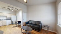 This deluxe efficiency studio is a thoughtfully organized single-floor unit that maximizes every inch of its compact footprint. The open-concept living area features warm hardwood-style flooring, neutral gray walls, and tall ceilings that lend a sense of openness. A large window allows natural light to fill the space, creating a bright and welcoming atmosphere. The living area comfortably accommodates a sofa and coffee table arrangement, while a ceiling fan adds practical comfort overhead.

The kitchen is well-equipped with light wood cabinetry, dark granite-style countertops, and a full suite of white appliances including a range with electric cooktop, over-the-range microwave, dishwasher, and a full-size refrigerator. Track lighting overhead ensures the kitchen is well-lit for everyday use. A half-wall partition subtly separates the kitchen from the sleeping area, which features carpeted flooring for added comfort.

The bathroom is clean and functional, complete with a tub-shower combination, a single vanity with matching cabinetry and dark countertop, an overhead storage cabinet, and hardwood-style flooring. The layout as a whole is efficient and cohesive, making it a practical and comfortable living solution.