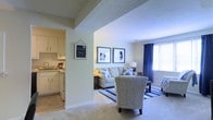 This single-floor residence offers a comfortable and inviting layout designed for easy everyday living. The living area serves as the heart of the home, featuring plush carpeting, large windows dressed with navy blue curtains that allow generous natural light to fill the room, and tasteful wall décor that adds a personal touch to the space. The seating arrangement, anchored by a striped area rug, creates a relaxed and well-organized gathering area with a warm, homey ambiance.

An open archway connects the living area to the kitchen, creating a seamless flow between spaces. The kitchen is outfitted with white cabinetry, tile flooring, and standard appliances, offering a clean and functional workspace. The light-colored walls and textured ceilings throughout contribute to a bright and airy atmosphere.

The neutral color palette of soft whites, beiges, and subtle blue accents ties the space together cohesively, giving it a calm and welcoming character. The single-floor layout ensures convenient accessibility, with all areas easily reachable without the need for stairs. Overall, this residence balances practicality with a cozy aesthetic, making it a comfortable place to call home.