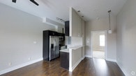This single-floor residence showcases a sleek, contemporary design with an open-concept layout that promotes a seamless flow between living spaces. The kitchen is a standout feature, equipped with stainless steel appliances including a French door refrigerator, microwave, and oven, all complemented by bold dark cabinetry that creates a striking contrast against the light neutral walls. A peninsula counter with pendant lighting above serves as a natural divider between the kitchen and the adjoining living area, offering both additional prep space and casual seating. Track lighting is installed to illuminate the kitchen workspace efficiently. Rich, dark hardwood flooring extends throughout the home, adding warmth and visual continuity to the modern aesthetic. The high ceilings amplify the sense of openness, making the space feel expansive and airy. A ceiling fan is visible, contributing to both comfort and style. A hallway leads to additional rooms, with natural light filtering in from a window at the far end, hinting at bright, well-lit private quarters. The overall ambiance is clean, modern, and functional — ideal for those who appreciate minimalist design paired with quality finishes.