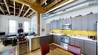Located at 300 Blackwell St in Durham, NC, this single-floor space masterfully combines industrial heritage with modern design. The open-concept layout is anchored by dramatic exposed timber beams and columns that span the ceiling alongside visible silver ductwork, creating a raw, warehouse-inspired aesthetic that pays homage to the building's industrial roots.

The kitchen is a standout feature, boasting sleek gray cabinetry with clean-lined hardware, a bold yellow tile backsplash that adds a vibrant pop of color, and a full suite of stainless steel appliances including a range, microwave, and dishwasher. A wine refrigerator is neatly integrated into the cabinetry, and a light-toned countertop provides ample workspace. The kitchen flows naturally into the dining and living areas, which are bathed in natural light from large windows.

Light hardwood flooring runs throughout, complementing the warmth of the timber beams above and grounding the industrial elements with an inviting feel. The living area is spacious and open, offering flexible furnishing options. The overall ambiance of this space is one of urban sophistication — where preserved architectural details meet thoughtfully curated modern finishes for a truly distinctive environment.